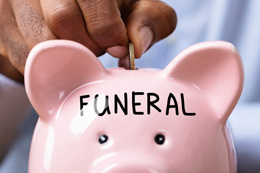 Person Saving Money For Funeral By Inserting Coin In Piggy Bank With Funeral Text